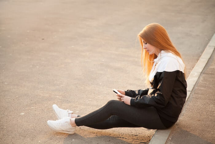A teen girl sitting on the sidewalk posting on social media and listening to music in Calgary. 