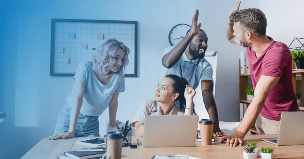 A cheerful, diverse group of four colleagues in a modern office celebrating a successful team effort. The team includes a woman with pink hair and three men, one of whom is high-fiving another. They are gathered around a work desk with laptops and coffee mugs